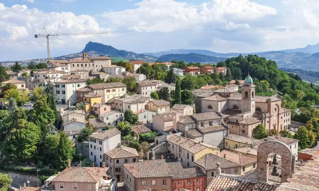 Panoramica di un borgo collinare con tetti rossi e una chiesa con campanile, incorniciato dal verde e dalle montagne. Un paesaggio che evoca la bellezza di Rimini e dintorni.
