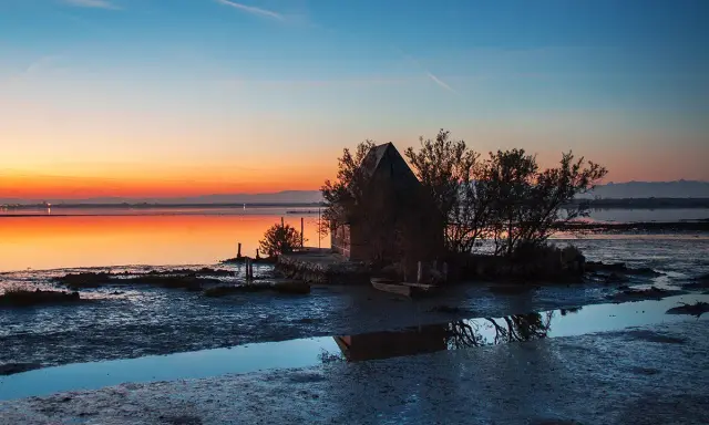 Tramonto sulla spiaggia di Marano, con una piccola capanna e un'imbarcazione riflessi sull'acqua calma.