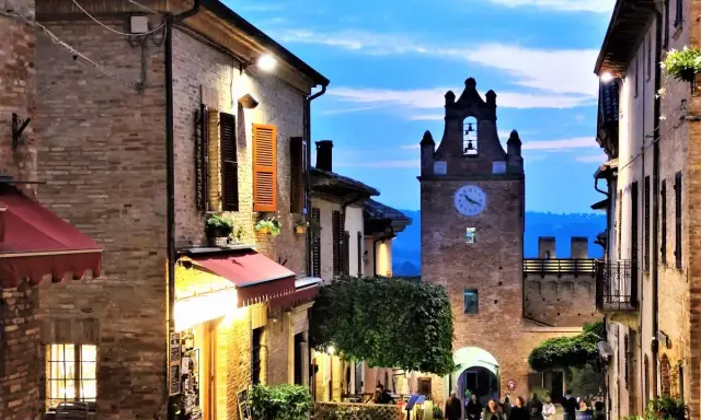 Serata magica al castello di Gradara. Vicoli illuminati, torre dell'orologio e gente che passeggia.