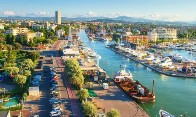 Vista aerea del porto di Rimini con barche ormeggiate, edifici e montagne sullo sfondo.