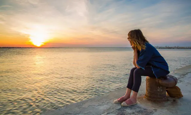 Donna seduta osserva il tramonto a Rimini, con il sole che si riflette sul mare calmo.
