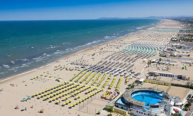 La riviera di Rimini in pieno sole, con un mare azzurro e una spiaggia affollatissima di ombrelloni colorati e bagnanti.
