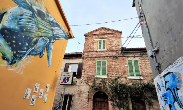 Un murale di balena colorata decora un edificio giallo a San Giuliano a Mare. Vicino, un edificio in mattoni con persiane verdi e un cartello di area pedonale.