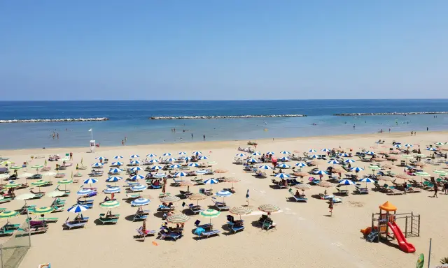 Spiaggia libera Viserba affollata di ombrelloni e lettini sotto un cielo azzurro. Bambini giocano nell'area attrezzata.