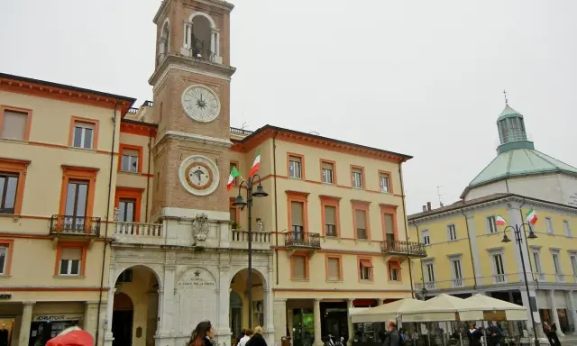 La piazza Rimini, con il suo imponente campanile e gli edifici storici, accoglie visitatori e residenti sotto un cielo nuvoloso.