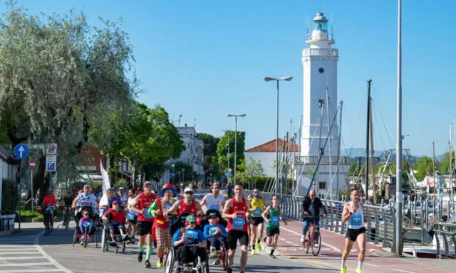 Atleti corrono lungo il percorso della Rimini Marathon, con il faro bianco e il porto sullo sfondo.