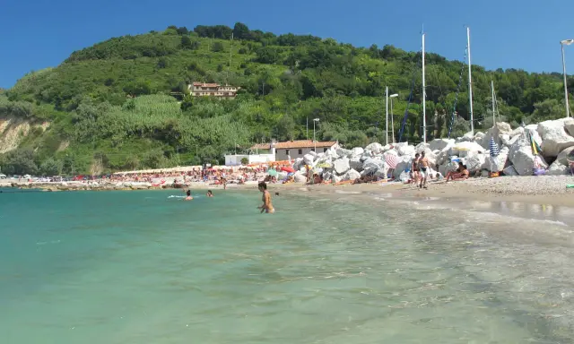 Spiaggia affollata con gente che nuota e prende il sole. Un promontorio verde domina la scena. Meglio Riccione o Gabicce Mare? Difficile scegliere!