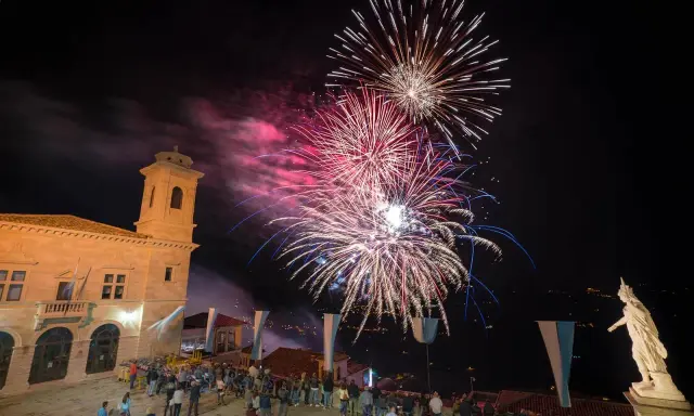 Spettacolo pirotecnico illumina il cielo notturno per la festa di San Marino. Folla ammira i fuochi d'artificio da una piazza con un edificio storico.