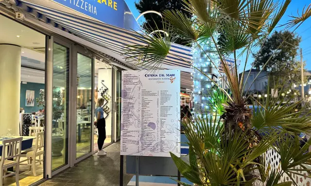 Osteria del Mare, ristorante pizzeria a Rimini centro, con menù esposto e tavoli all'esterno. Palme illuminate con lucine.