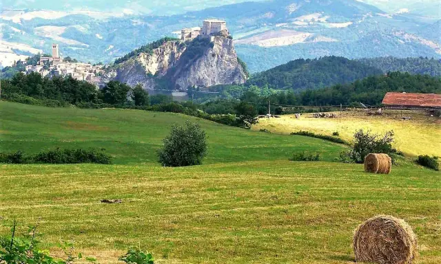 Campagna con borghi vicino Rimini, colline verdi, balle di fieno e un castello arroccato su uno sperone roccioso.
