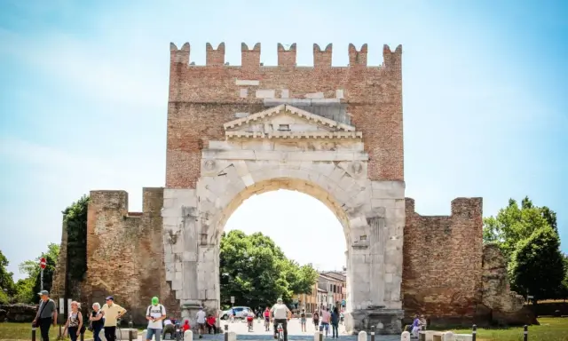 L'imponente arco d'augusto rimini, con le sue antiche pietre e la torre in mattoni, fa da sfondo a una vivace piazza con persone che passeggiano e ciclisti.