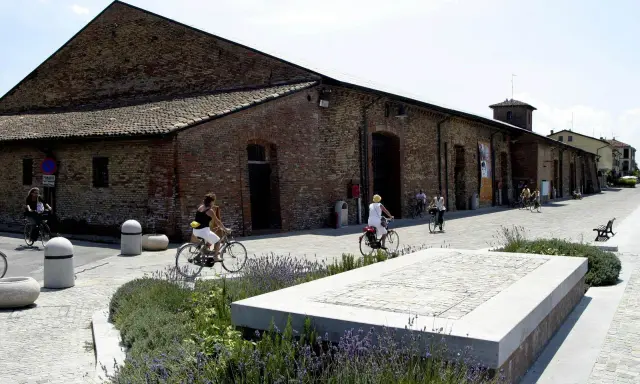 Passeggiata in bicicletta davanti ai Magazzini del Sale di Cervia, con persone che pedalano su un lungomare acciottolato, tra aiuole fiorite e antichi edifici in mattoni.