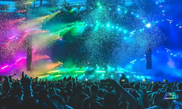 Folla in delirio a un concerto a Rimini, con coriandoli colorati e luci psichedeliche che illuminano la scena.