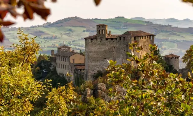 Castello medievale su una collina, circondato da alberi con foglie autunnali dai colori caldi. Il paesaggio collinare sullo sfondo evoca un senso di pace in questo mare autunno.