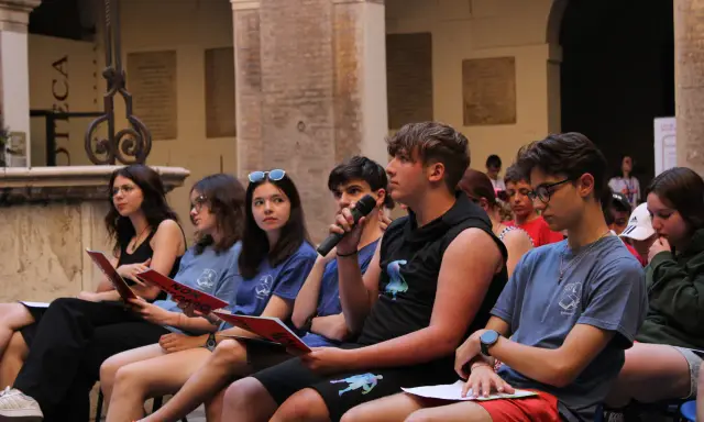 Giovani ascoltano un discorso, alcuni con fogli rossi in mano, durante un evento culturale a Rimini. Un ragazzo tiene un microfono.