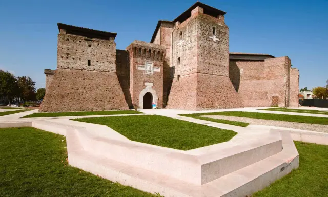 Imponente Castel Sismondo, con le sue torri in mattoni rossi e il cortile verdeggiante, si erge maestoso sotto un cielo azzurro.