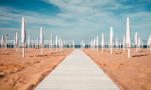 Passerella bianca su sabbia dorata, file di ombrelloni chiusi e strutture bianche sotto un cielo azzurro. Un'immagine serena del mare in Emilia-Romagna.