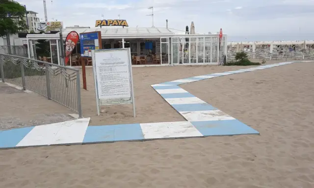 Percorso a strisce bianche e blu sulla sabbia del piazzale San Martino a Riccione, conduce al bar Papaya e alla spiaggia attrezzata.