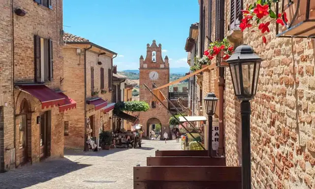 Un vicolo acciottolato a Gradara, con edifici in mattoni, fiori rossi e un campanile con orologio. Perfetto per scoprire cosa vedere.