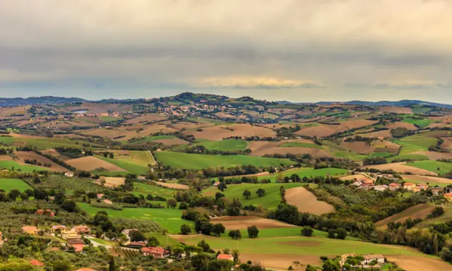 Dolci colline verdi e marroni della Valconca, Rimini, punteggiate da case e alberi, sotto un cielo nuvoloso.
