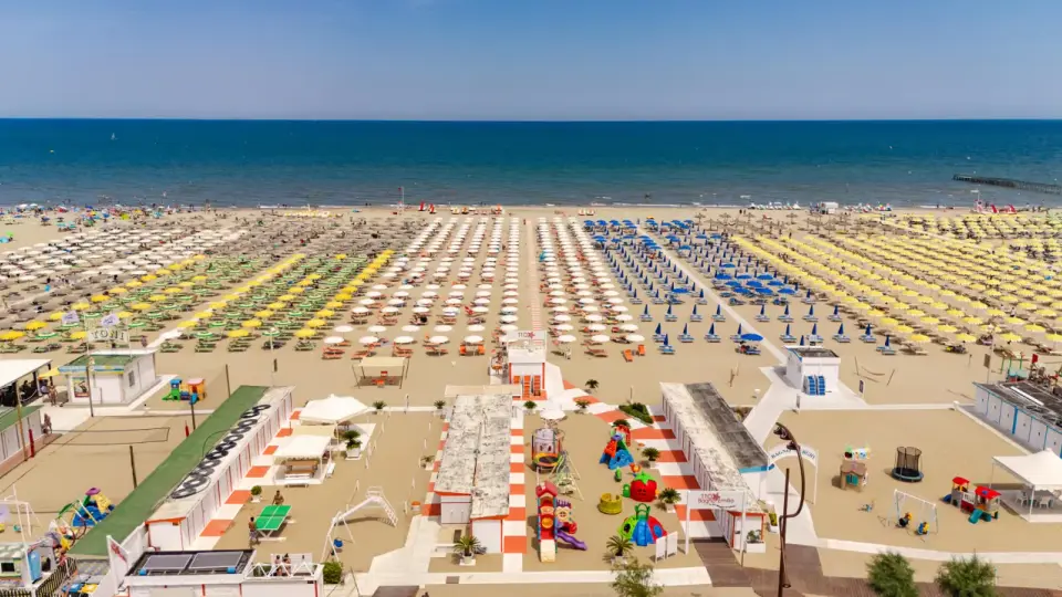 Ampia spiaggia di Rimini Rivazzurra con file ordinate di ombrelloni colorati, lettini, giochi per bambini e il mare azzurro all'orizzonte.