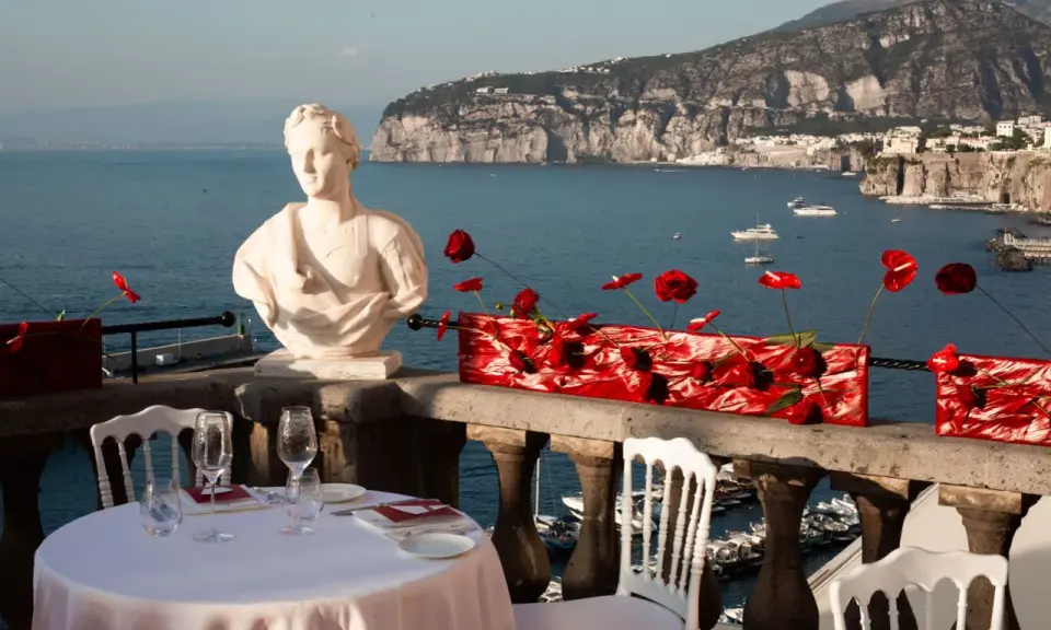 Tavolo apparecchiato con vista mare, perfetto per idee per Ferragosto. Busto in marmo, fiori rossi e panorama mozzafiato.