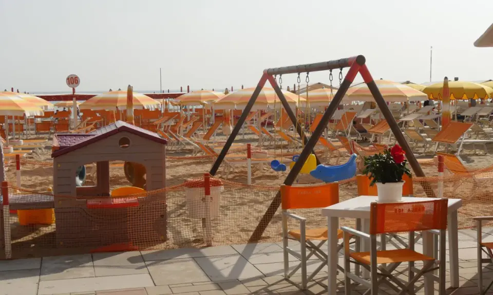 Area giochi e lettini sulla spiaggia 106 di Riccione, con ombrelloni arancioni e bianchi, altalena e casetta per bambini.