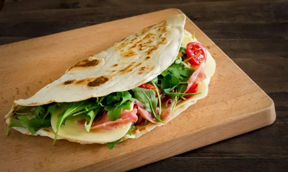 Piadina romagnola, un classico dal sapore autentico, ripiena di prosciutto, formaggio, rucola e pomodorini.