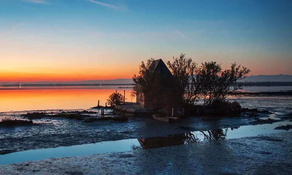 Tramonto sulla spiaggia di Marano, con una piccola capanna e un'imbarcazione riflessi sull'acqua calma.