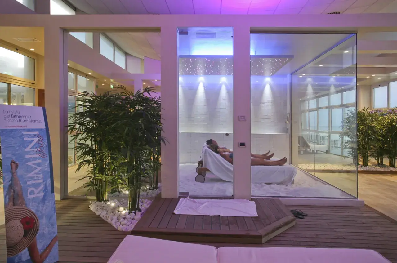 Un'oasi talassoterapico a Rimini, con due persone che si rilassano su lettini in un ambiente moderno e luminoso, circondato da piante di bambù e illuminazione soffusa.