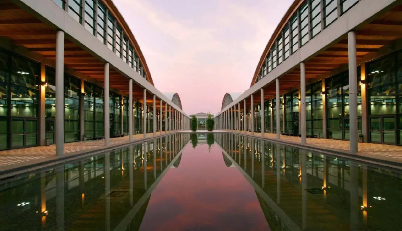Ingresso moderno della fiera a Rimini, con un lungo specchio d'acqua che riflette le arcate e il cielo rosa del tramonto.