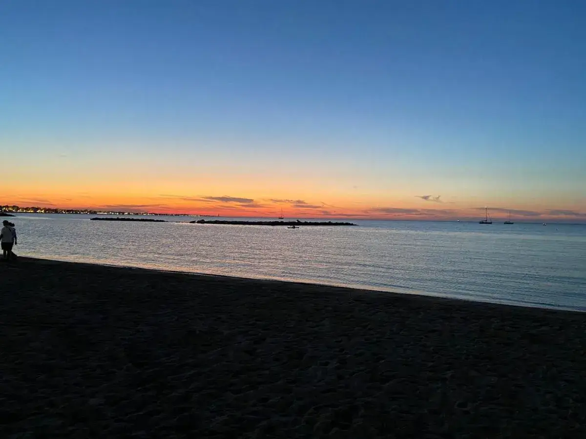 Tramonto sulla spiaggia di Rimini, con il cielo che sfuma dall'arancione al blu. Due persone passeggiano sulla sabbia scura, mentre in mare si vedono barche a vela.