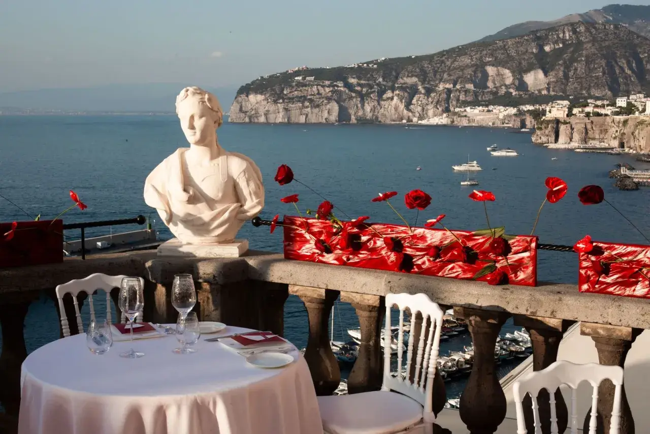Tavolo apparecchiato con vista mare, perfetto per idee per Ferragosto. Busto in marmo, fiori rossi e panorama mozzafiato.