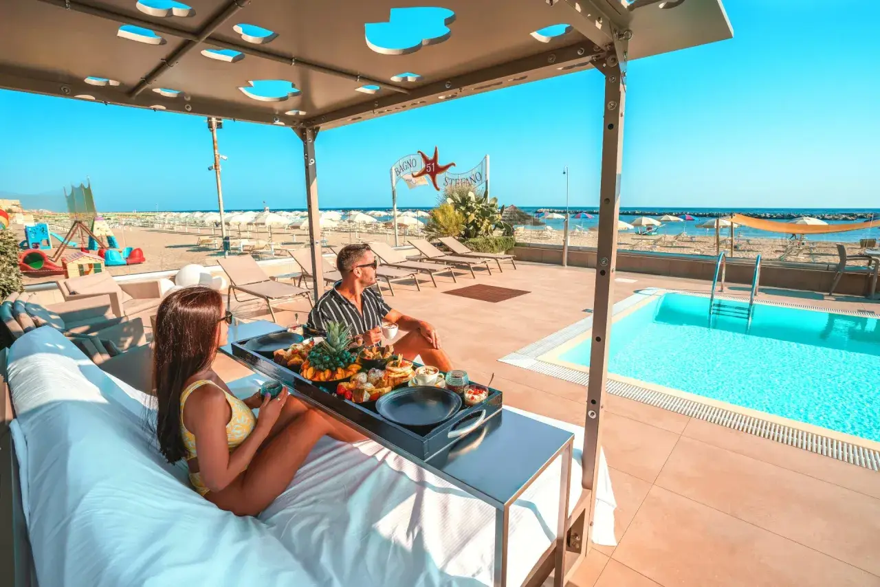 Coppia rilassata su lettini con vassoio di frutta e caffè, con piscina e spiaggia di Rimini sullo sfondo.