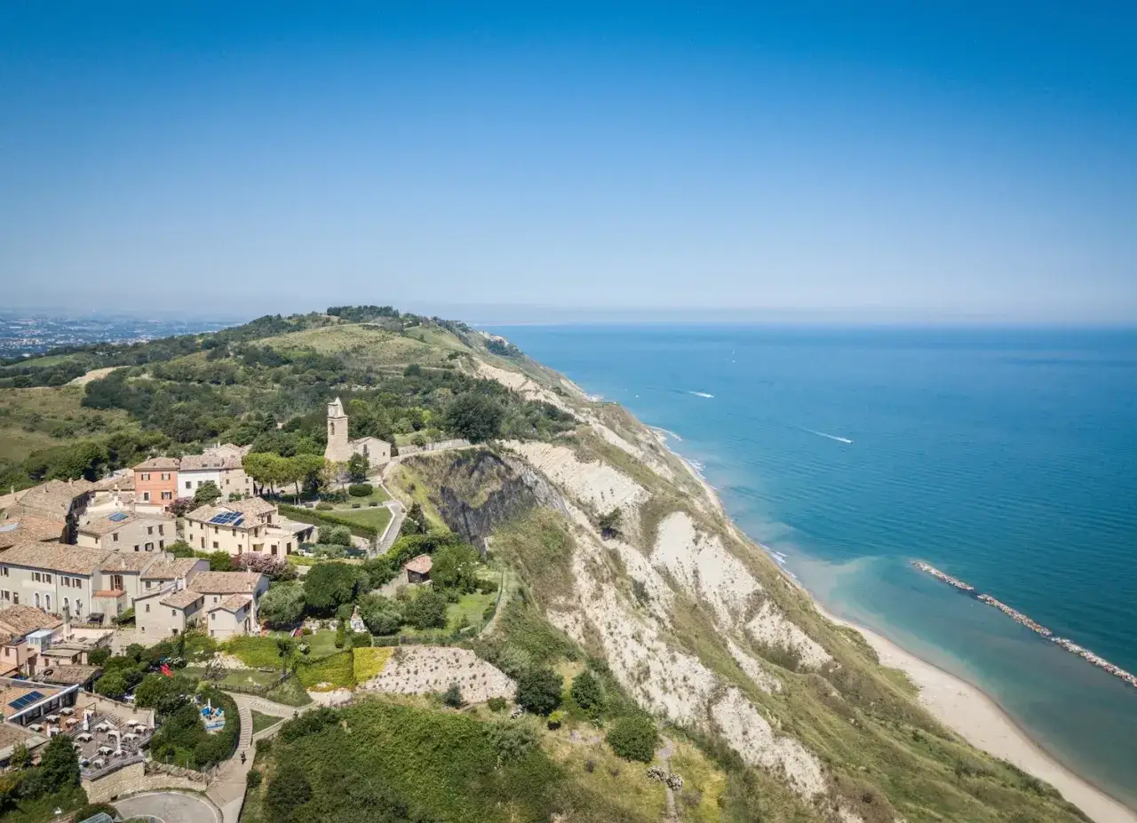 Vista aerea di un borgo collinare affacciato sul mare Adriatico. Le case colorate e la chiesa si stagliano sul verde, con scogliere a picco sull'acqua. Un luogo che ispira tante belle gabicce mare recensioni.