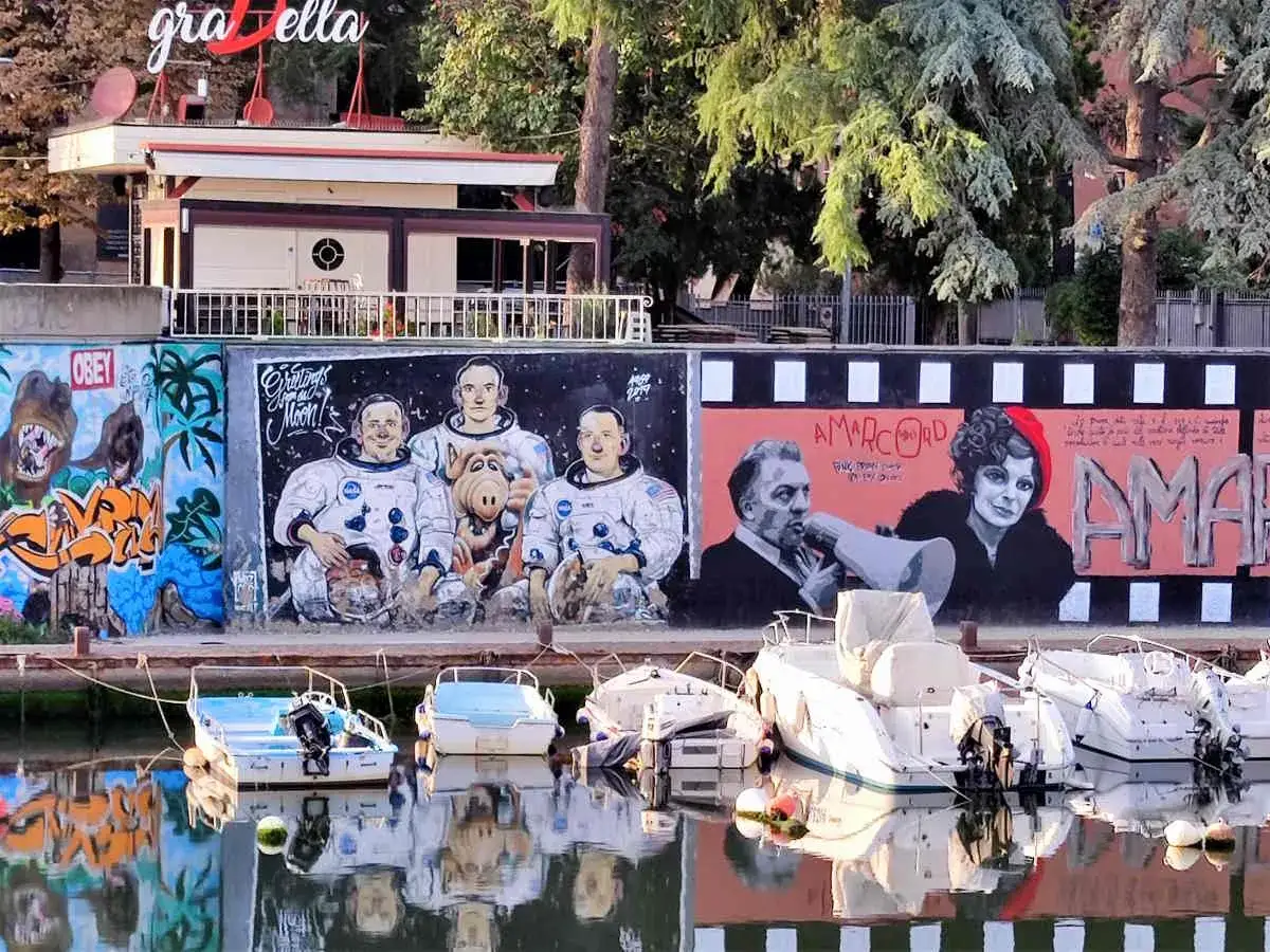Graffiti artistici a Rimini Borgo San Giuliano: astronauti con Alf, un uomo con megafono e una donna con berretto rosso. Barche ormeggiate sull'acqua.