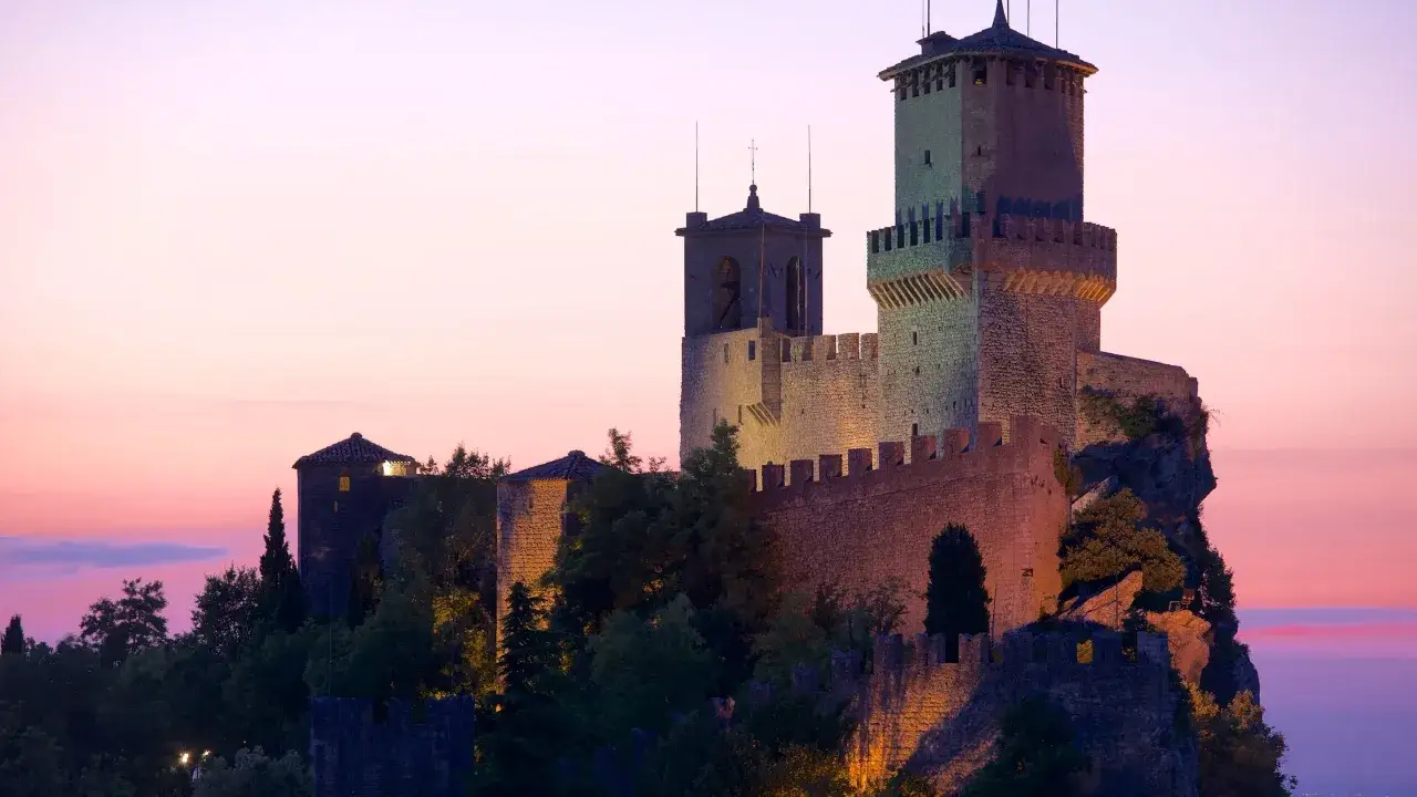 Torri illuminate di San Marino città al tramonto, con un cielo rosa e viola.
