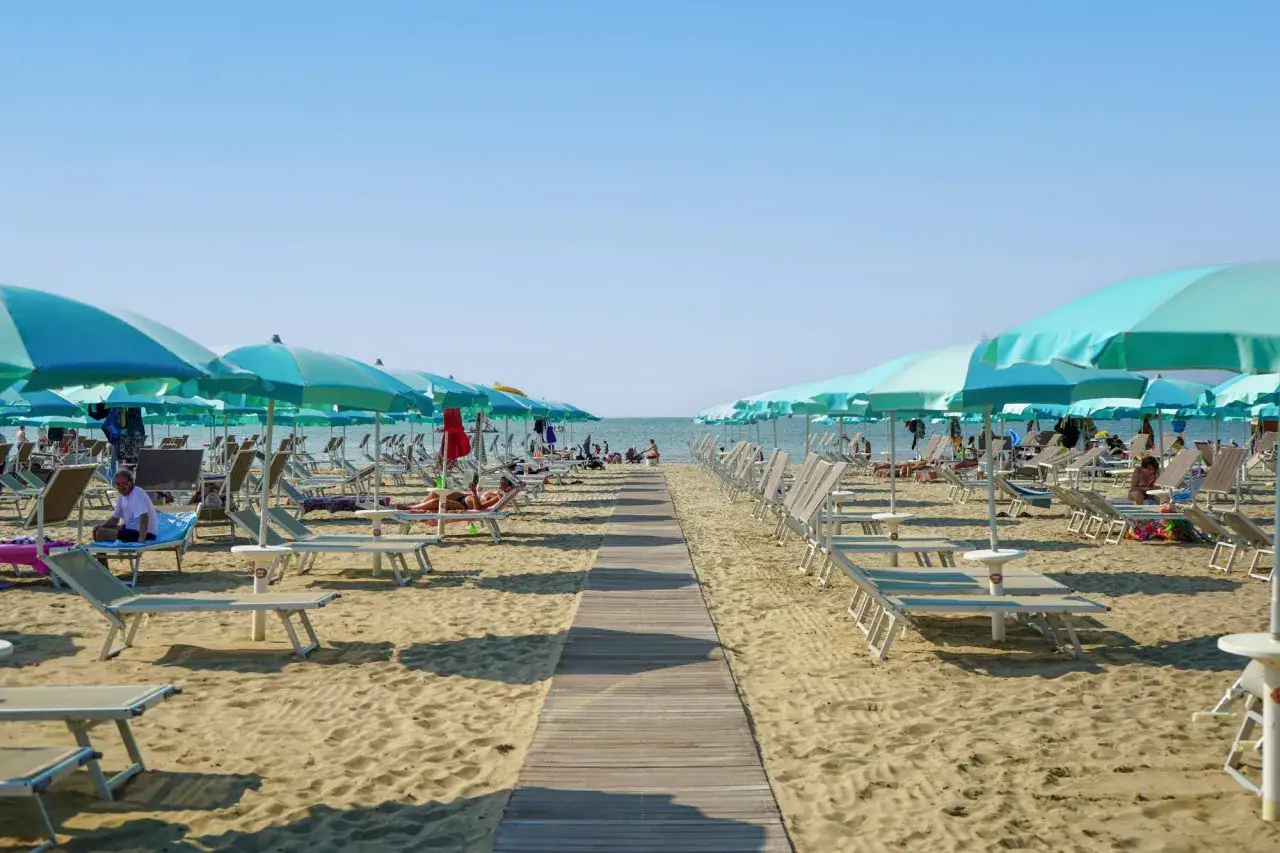 Un percorso in legno conduce al mare di Rimini, fiancheggiato da file di lettini e ombrelloni azzurri sotto un cielo limpido.