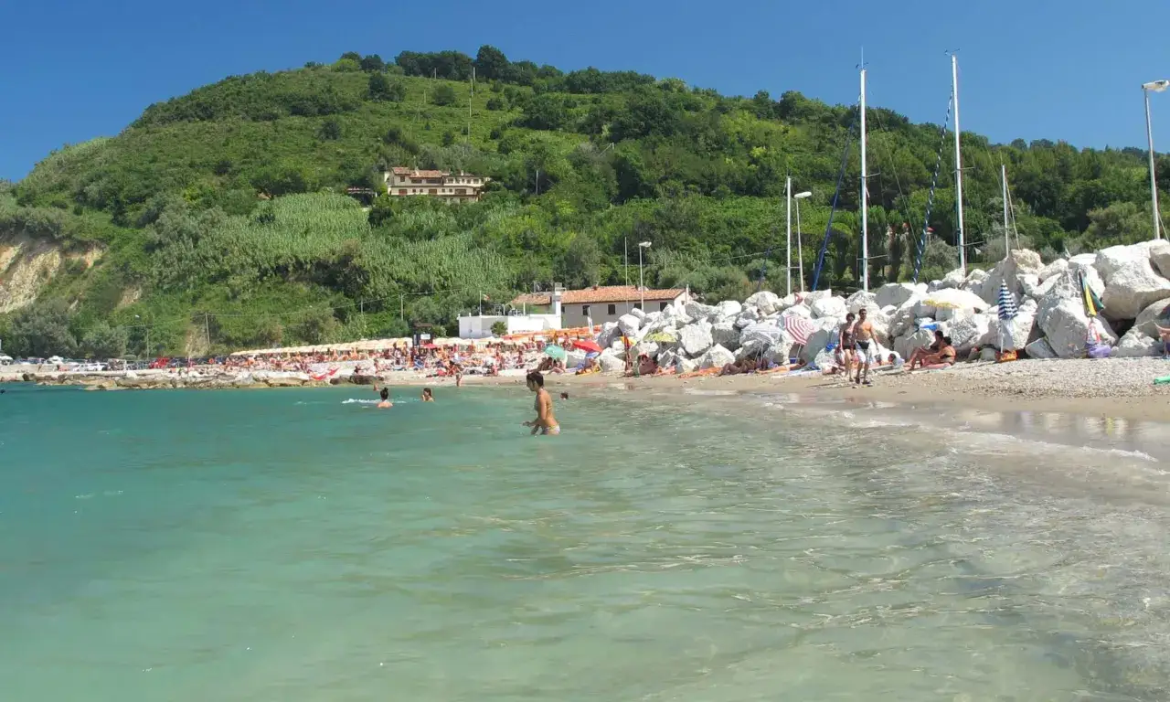Spiaggia affollata con gente che nuota e prende il sole. La collina verdeggiante sullo sfondo fa pensare a un dilemma: meglio Riccione o Gabicce Mare?