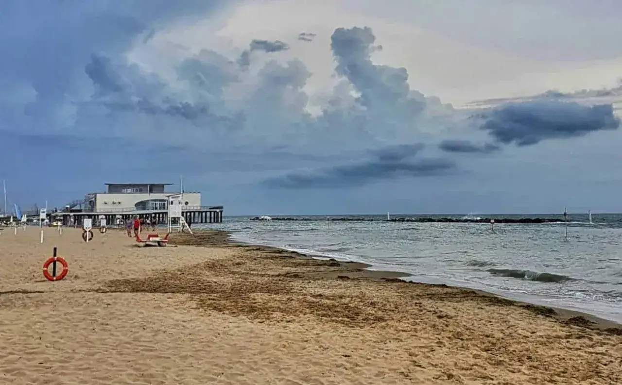 Spiaggia deserta con un edificio bianco su palafitte, onde che lambiscono la riva e nuvole minacciose. Un'atmosfera da mare d'inverno.