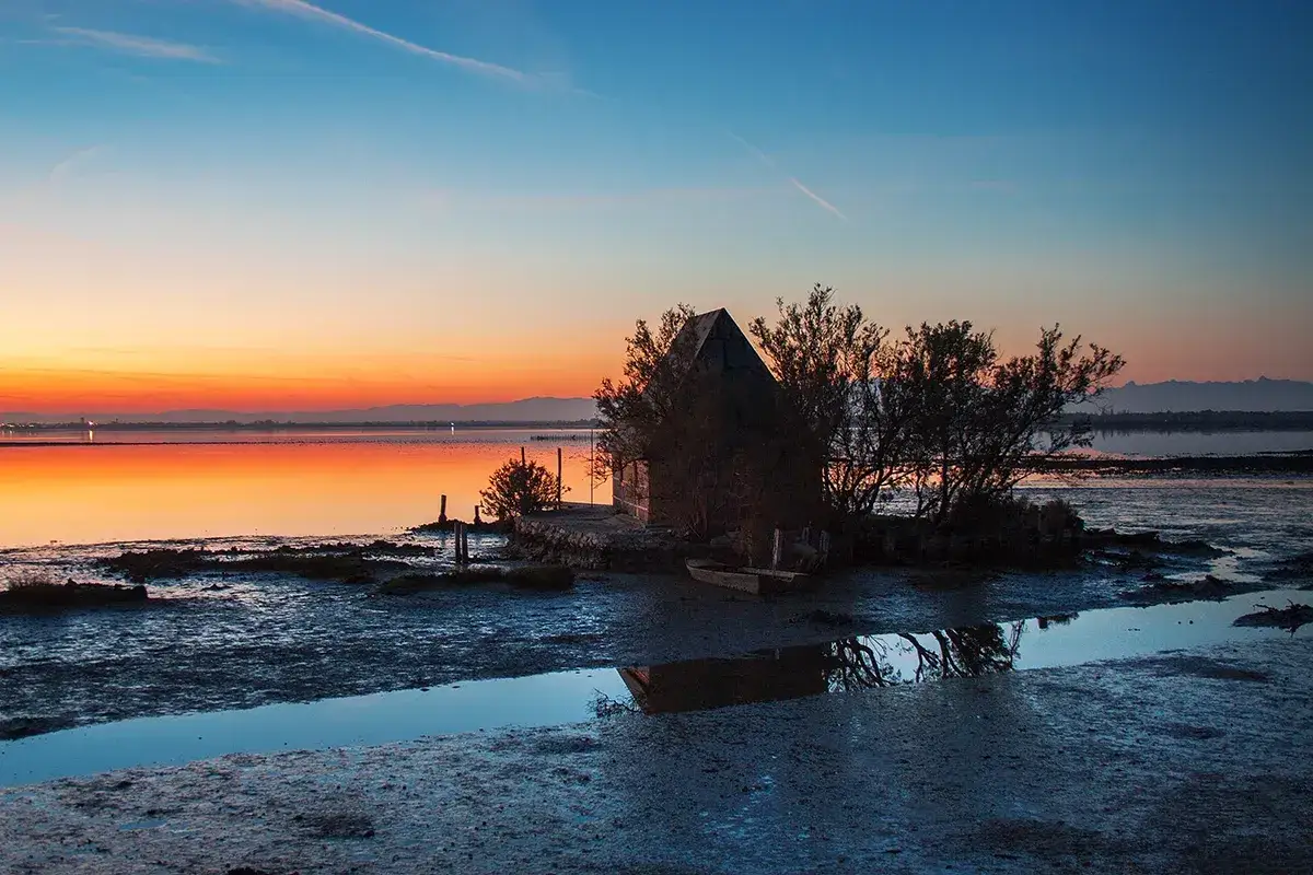 Tramonto sulla spiaggia di Marano, con un piccolo capanno e un'imbarcazione riflessi sull'acqua.