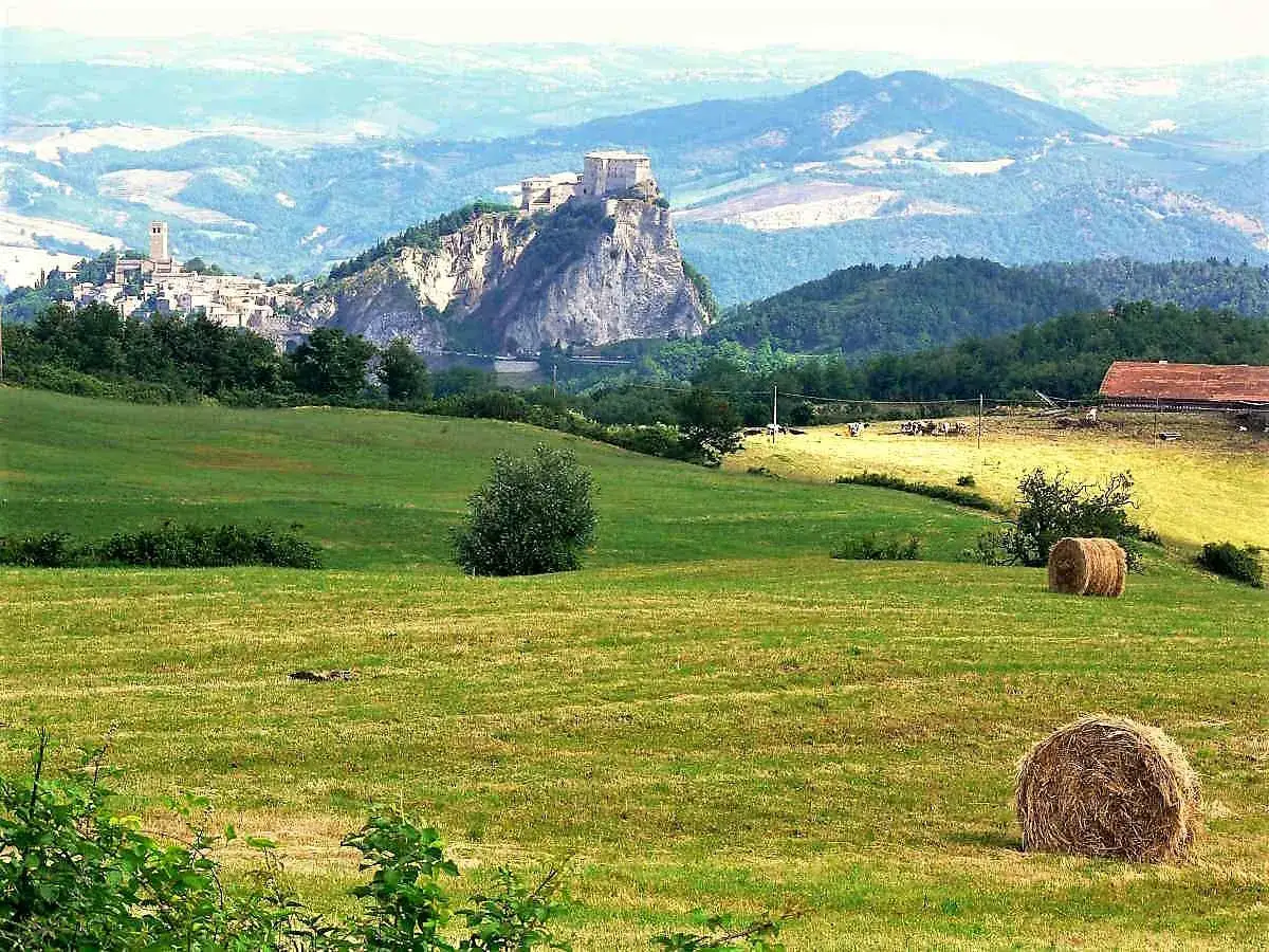 Campagna collinare con borgo arroccato e castello, ideale per trekking rimini. Balze verdi, balle di fieno e un casolare.