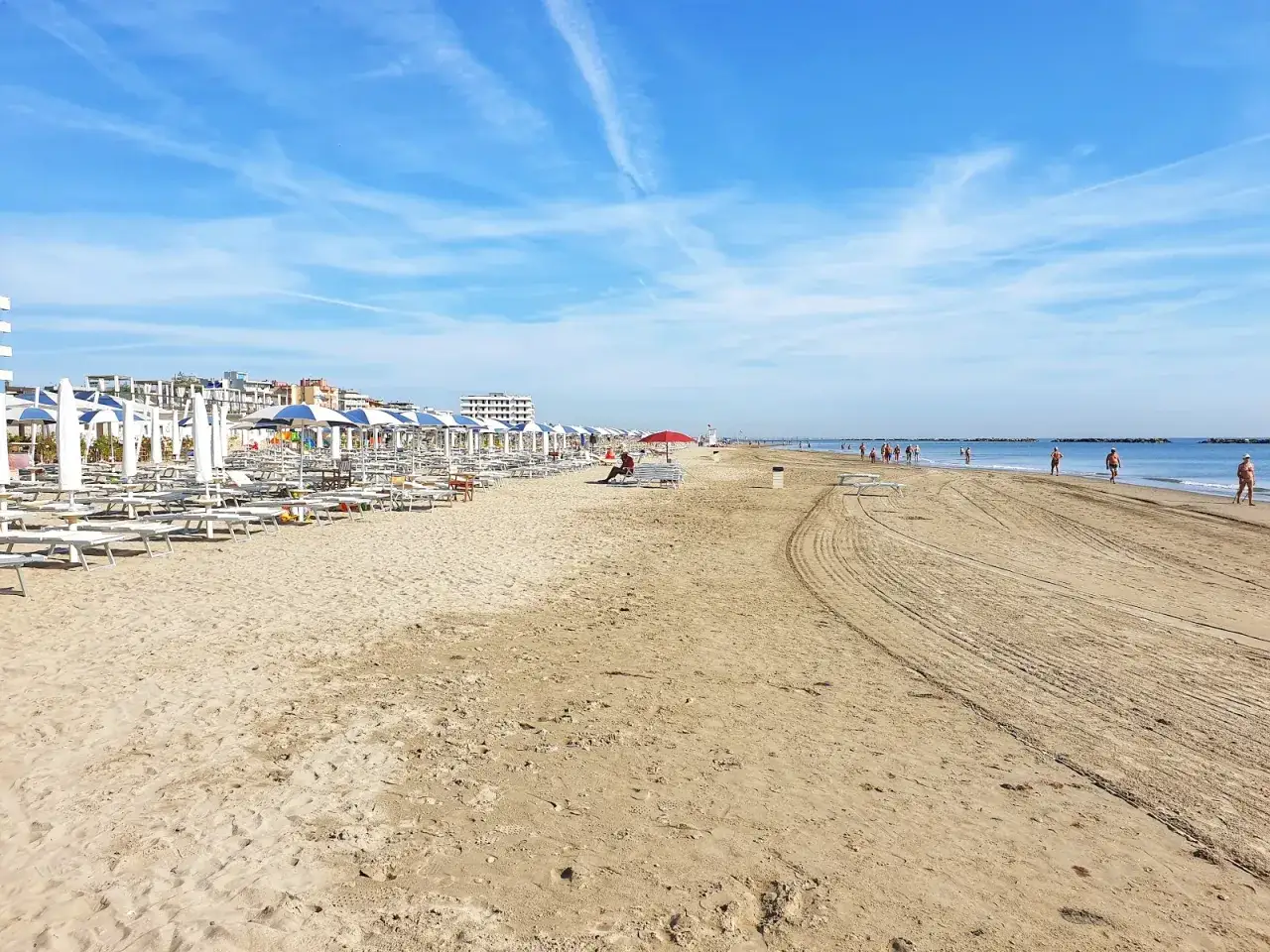 Spiaggia di Lido Adriano con file di ombrelloni e lettini, bagnanti che passeggiano sul bagnasciuga sotto un cielo azzurro.