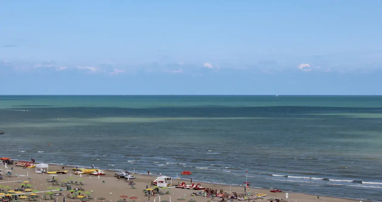 Spiaggia affollata a Rivazzurra, con ombrelloni, lettini, pedalò e il mare azzurro all'orizzonte.