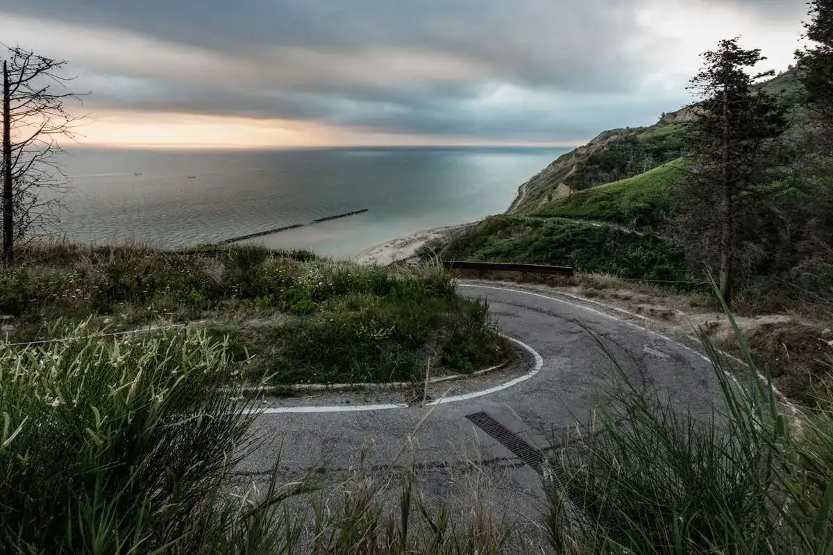 Strada panoramica a tornanti verso Gabicce Mare, con il mare Adriatico e la costa verdeggiante sotto un cielo nuvoloso.