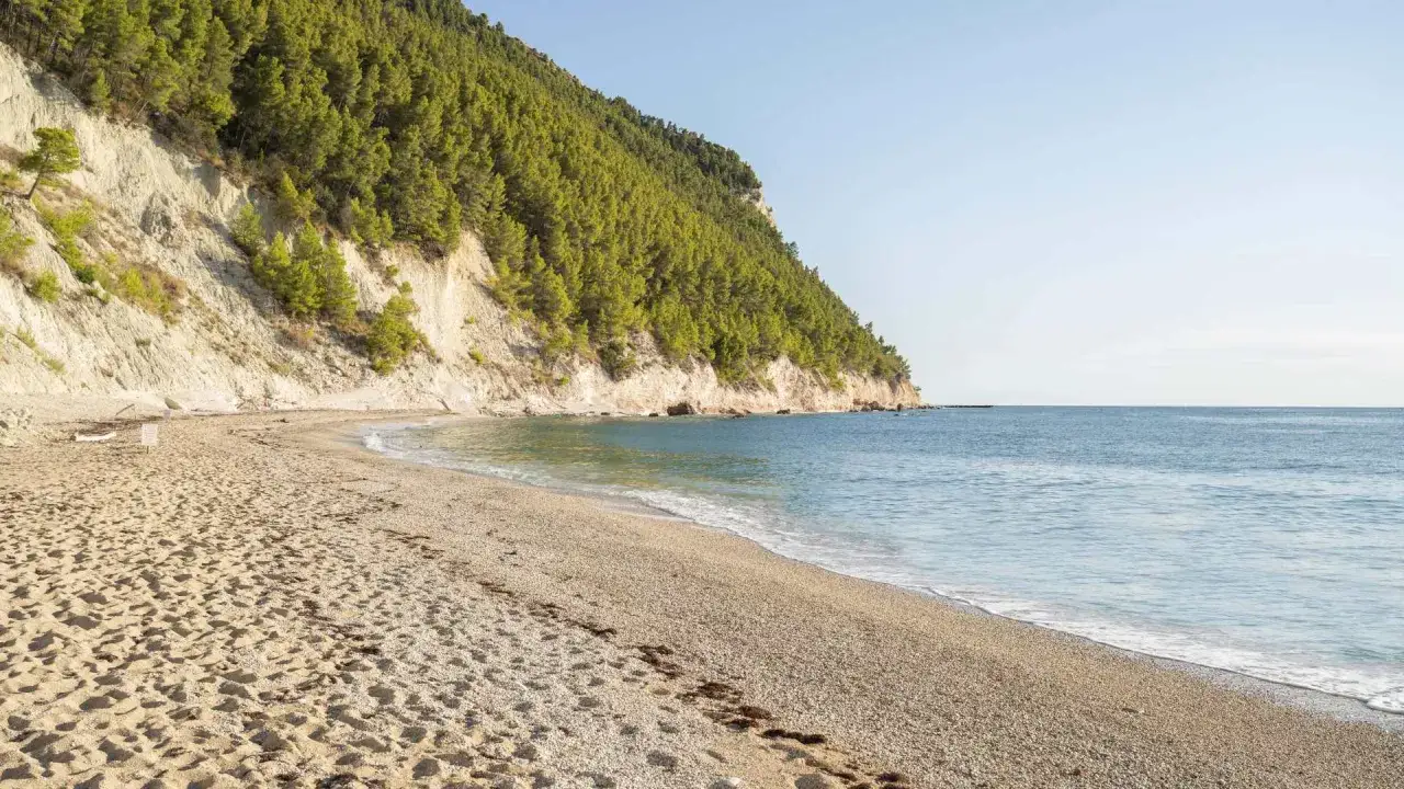 La spiaggia di Castelmezzano si estende serena, con sabbia dorata e ciottoli levigati, lambita da un mare azzurro e calmo sotto una scogliera verdeggiante.