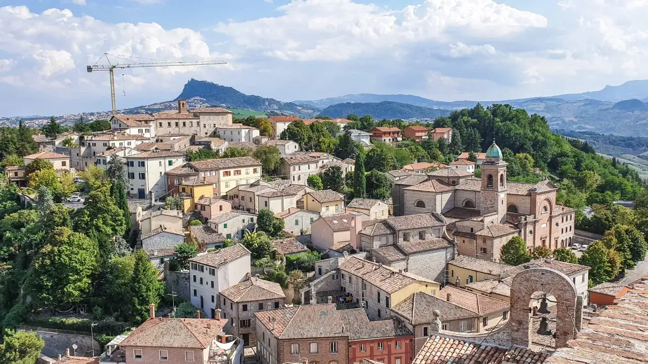 Panoramica di un borgo collinare con case dai tetti rossi, una chiesa con campanile e montagne sullo sfondo. Un paesaggio incantevole nei dintorni di Rimini.