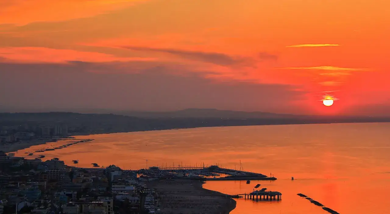 Tramonto infuocato sul centro storico e lungomare di Gabicce Mare. Il sole cala sul mare, tingendo il cielo e l'acqua di arancione e rosso.