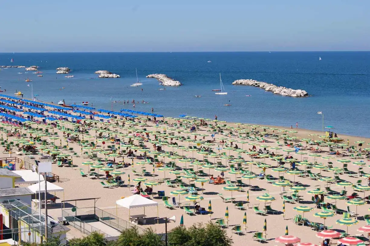 Ampia spiaggia libera a Viserba, con file ordinate di ombrelloni verdi e gialli, lettini e persone che si godono il sole. Il mare azzurro e calmo invita a un bagno rinfrescante.