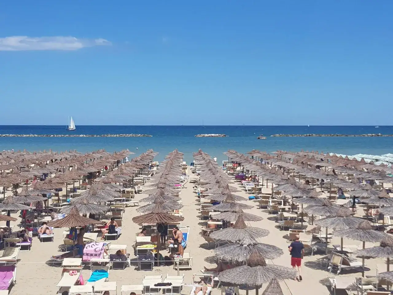 Un affollato bagno cesenatico con file di ombrelloni di paglia e lettini sulla sabbia dorata. Il mare azzurro e un veliero all'orizzonte.